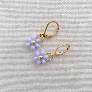 May include: A pair of gold-colored leverback earrings, each featuring a light purple flower with a gold-colored center. The earrings are displayed on a neutral-colored fabric background.