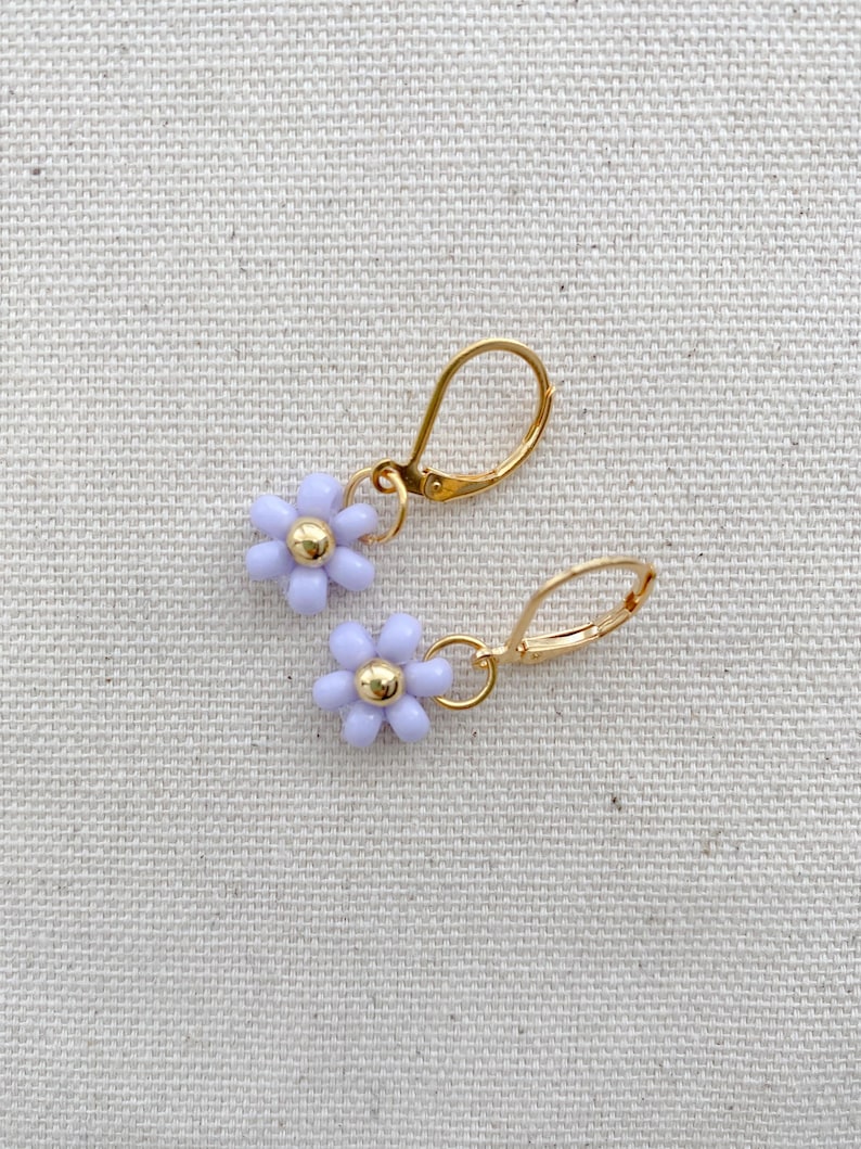 May include: A pair of gold-colored leverback earrings, each featuring a light purple flower with a gold-colored center. The earrings are displayed on a neutral-colored fabric background.
