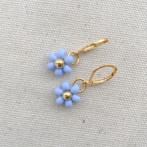 May include: A pair of gold-colored leverback earrings, each featuring a light blue daisy with a gold-colored center. The earrings are displayed on a neutral-colored fabric background. The daisy flower earrings are a delicate and feminine accessory.