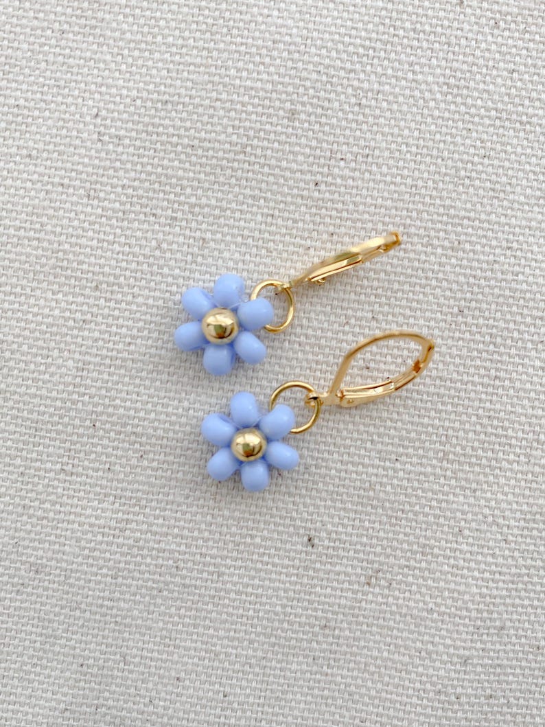 May include: A pair of gold-colored leverback earrings, each featuring a light blue daisy with a gold-colored center. The earrings are displayed on a neutral-colored fabric background. The daisy flower earrings are a delicate and feminine accessory.