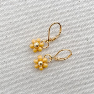 May include: A pair of gold-colored leverback earrings. Each earring features a small, yellow daisy flower with a gold-colored center. The earrings are displayed on a textured, off-white surface.
