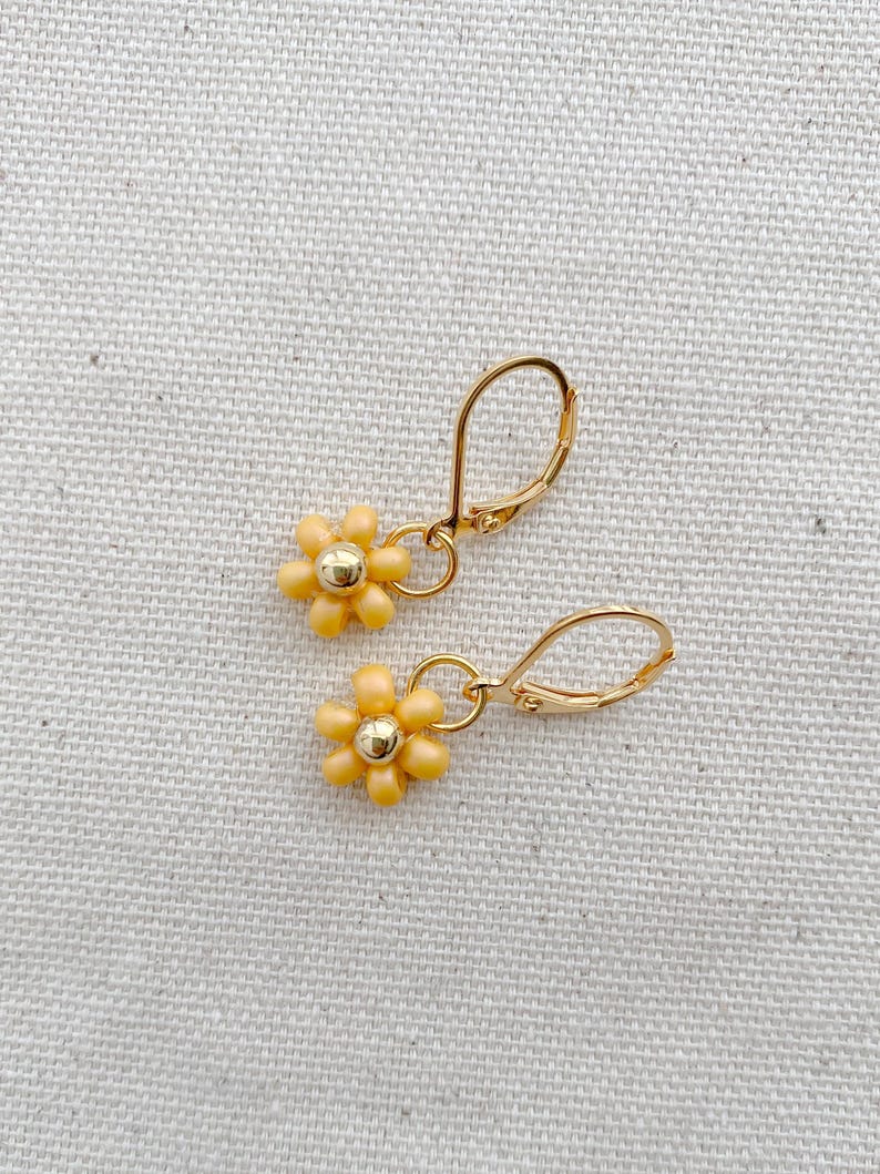May include: A pair of gold-colored leverback earrings. Each earring features a small, yellow daisy flower with a gold-colored center. The earrings are displayed on a textured, off-white surface.