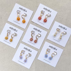 May include: A collection of beaded flower earrings in various colors, including orange, yellow, pink, white, and blue. Each pair is displayed on a white card with the brand name "mimita" printed on it. The earrings have gold-colored hoops.