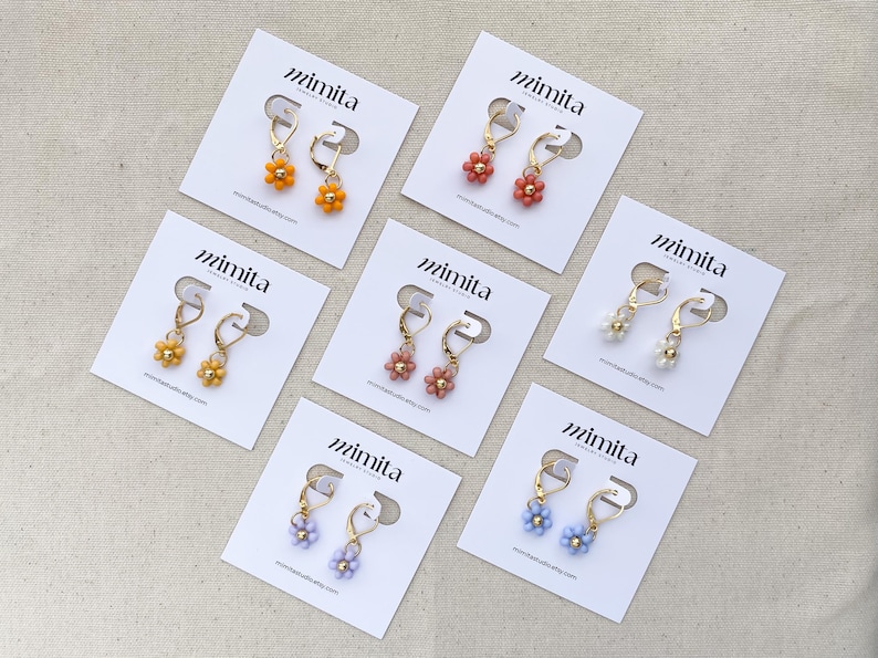 May include: A collection of beaded flower earrings in various colors, including orange, yellow, pink, white, and blue. Each pair is displayed on a white card with the brand name "mimita" printed on it. The earrings have gold-colored hoops.