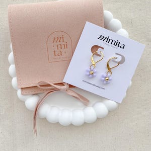 May include: A pair of gold-colored dangle earrings with small, light purple flower-shaped beads. The earrings are displayed on a white card with the text "mimita jewelry studio" and a website address. A light pink pouch and a white decorative tray are also in the image.