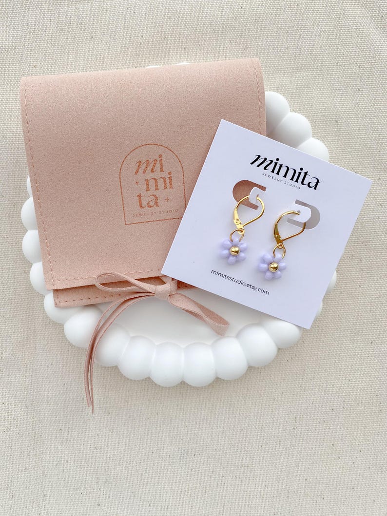 May include: A pair of gold-colored dangle earrings with small, light purple flower-shaped beads. The earrings are displayed on a white card with the text "mimita jewelry studio" and a website address. A light pink pouch and a white decorative tray are also in the image.