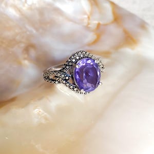 May include: A silver ring with a large, oval-cut purple gemstone. The ring's band features intricate detailing with smaller, clear stones. The ring is displayed on a white and beige seashell, creating a contrast of textures and colors.