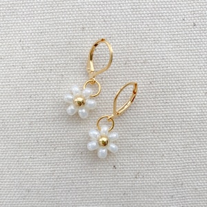May include: A pair of gold-colored hoop earrings, each featuring a small white daisy pendant with a gold-colored center. The earrings are displayed on a textured, off-white surface.
