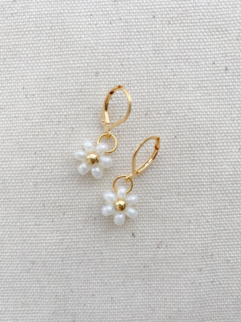 May include: A pair of gold-colored hoop earrings, each featuring a small white daisy pendant with a gold-colored center. The earrings are displayed on a textured, off-white surface.