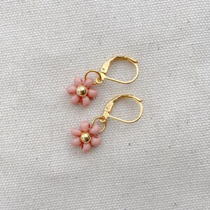 May include: A pair of gold-colored leverback earrings, each featuring a small, pink beaded flower with a gold-colored center. The earrings are displayed on a textured, off-white surface.