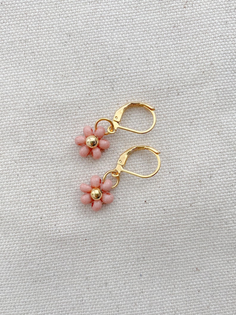 May include: A pair of gold-colored leverback earrings, each featuring a small, pink beaded flower with a gold-colored center. The earrings are displayed on a textured, off-white surface.