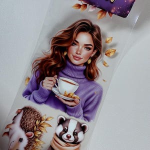 Może przedstawiać: Dekoracyjna naklejka przedstawiająca kobietę w fioletowym golfie trzymającą filiżankę herbaty. Naklejka zawiera również ilustracje jeża, borsuka i jesiennych liści. Ogólny motyw to przytulna jesień.