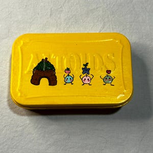 May include: A rectangular yellow tin with a decorative design. The tin features a brown and green illustration of a small hut and three cartoon characters with colorful details. The word "ANTIOUDS" is printed on the tin.