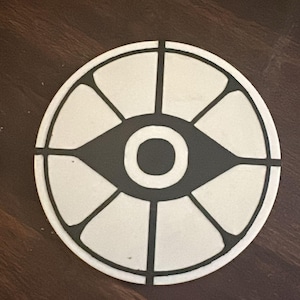 Puede incluir: Un posavasos redondo y blanco con un diseño de ojo negro en el centro. El ojo está rodeado por un contorno negro y segmentado por líneas negras. El posavasos está sobre una superficie de madera marrón oscuro.
