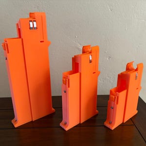 May include: Three bright orange, rectangular objects of varying sizes are arranged on a dark wooden surface. Each object has a unique shape with a series of cutouts and metal accents. The objects appear to be 3D-printed or manufactured.
