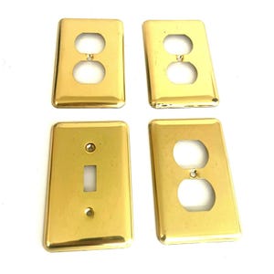 May include: A set of four rectangular wall plates with a polished brass finish. Three plates feature a standard duplex outlet configuration with two circular openings, and one plate features a single rectangular toggle switch opening.