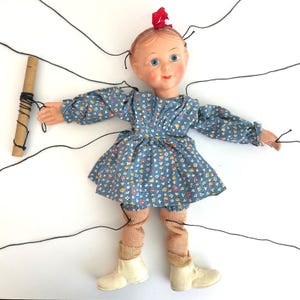 May include: Vintage marionette doll with a blue floral dress, tan socks, and white boots. The doll has a painted face with blue eyes and a red bow in her hair. Black strings are attached to the doll for manipulation, and a cardboard control is in the doll's left hand.