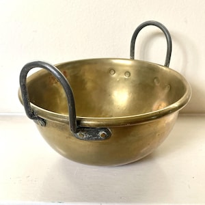 May include: A vintage brass bowl with two arched, dark metal handles. The bowl has a rounded shape and a polished, golden finish. Rivets secure the handles to the bowl's sides. The interior of the bowl is visible.
