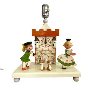 May include: A vintage wooden lamp featuring a clock tower design with a clock face showing the time. The lamp includes two figurines and two small rabbits. The clock tower is orange and white, and the figurines are dressed in green and white outfits.