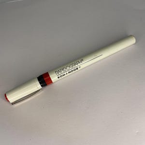 May include: A white Rapidograph Koh-I-Noor pen with a red tip and black band. The pen has a silver clip and the brand name is printed in black. The pen is designed for technical drawing and writing.