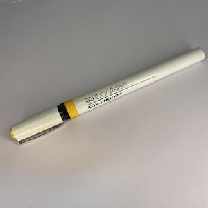 May include: A white Rapidograph technical pen with a yellow accent and a silver clip. The pen has black text that reads "RAPIDOGRAPH® KOH-I-NOOR®". The pen is designed for precision drawing and drafting.