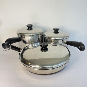 May include: A set of stainless steel cookware, including a frying pan with a lid, a saucepan with a handle, and two pots with lids. The lids have black knobs, and the saucepan and frying pan have black handles.