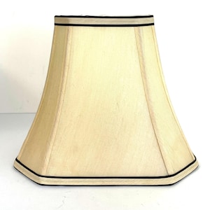 May include: A bell-shaped lampshade with a cream-colored fabric exterior. The lampshade features a structured, flared design with distinct vertical seams and a thin, dark black trim accenting both the top and bottom edges of the piece.