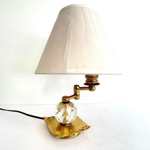 May include: A vintage brass desk lamp with a cream-colored lampshade. The lamp features a faceted crystal ball and an adjustable arm. The base is a square brass plate. A black power cord extends from the base.