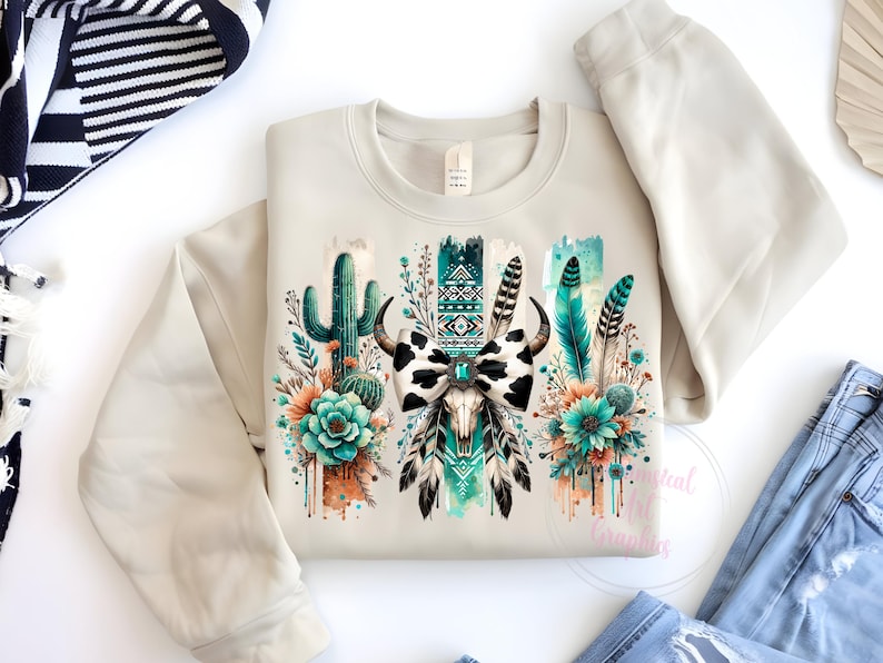 May include: A beige sweatshirt featuring a graphic print of a cow skull with a bow, surrounded by cacti, feathers, and flowers in shades of turquoise, green, and brown.