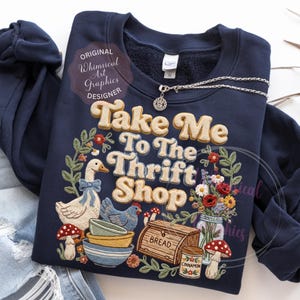 Puede incluir: Sudadera azul marino con el texto "Take Me To The Thrift Shop" en una fuente retro. El dise&ntilde;o incluye ilustraciones de un ganso, una gallina, setas, cuencos, pan y flores. Una cadena de plata con un colgante est&aacute; encima.