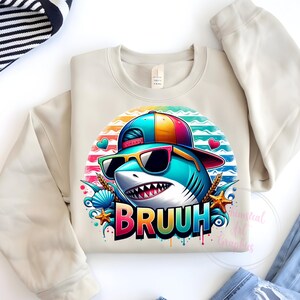 Puede incluir: Sudadera beige de cuello redondo con un tibur&oacute;n de dibujos animados con gafas de sol y un sombrero colorido. El gr&aacute;fico del tibur&oacute;n incluye la palabra "BRUUH" en una fuente de arco&iacute;ris, rodeada de corazones, conchas marinas y estrellas de mar.