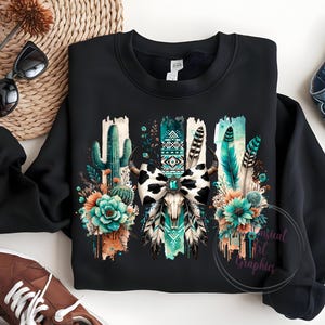 May include: Black sweatshirt featuring a graphic print of a cow skull with a turquoise and black bow, surrounded by cacti, feathers, and flowers. The design has a watercolour effect.