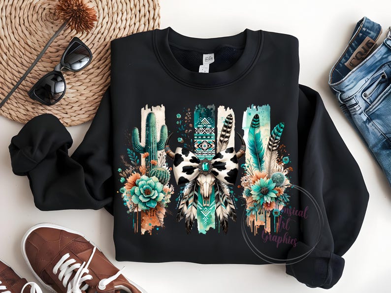 May include: Black sweatshirt featuring a graphic print of a cow skull with a turquoise and black bow, surrounded by cacti, feathers, and flowers. The design has a watercolour effect.