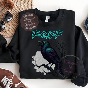 May include: Black sweatshirt with a raven illustration and the teal text "F-CAW-F". The design includes a tree silhouette and a moon shape. The sweatshirt has a round neck. The design is by Whimsical Art Graphics.