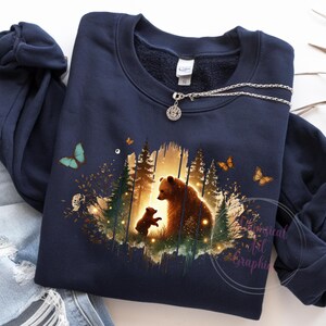 May include: A navy blue sweatshirt with a graphic print of a mother bear and her cub standing in a forest setting. The graphic is surrounded by a watercolor-like effect and features butterflies.