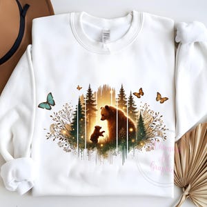 May include: White sweatshirt with a graphic print of a mother bear and her cub standing in a forest with butterflies. The graphic is in shades of brown, green, and gold.