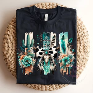 May include: Black t-shirt featuring a graphic print of a cow skull adorned with a turquoise and white bow, surrounded by cactus, feathers, and flowers. The design has a southwestern theme.