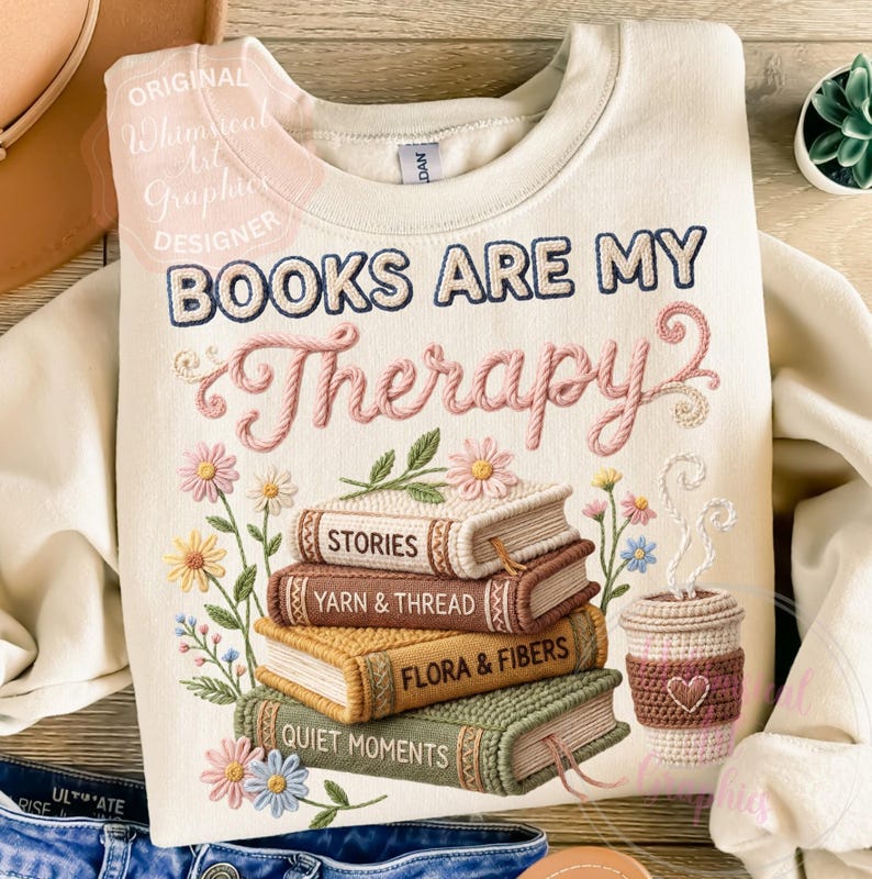 May include: Cream-colored sweatshirt with the embroidered text "BOOKS ARE MY Therapy". Below the text is a stack of books with the titles "STORIES", "YARN & THREAD", "FLORA & FIBERS", and "QUIET MOMENTS". A crocheted coffee cup is on the right.