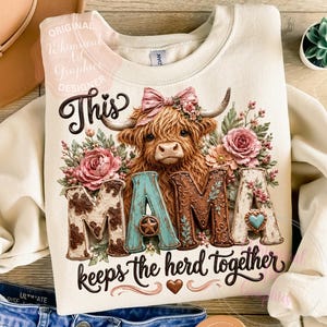 May include: A cream-coloured sweatshirt featuring a Highland cow graphic and the text "This MAMA keeps the herd together." The word "MAMA" is in a western-style font with floral and cowhide patterns. The cow has a pink bow and floral accents.