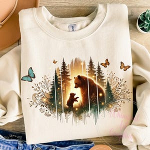 May include: A beige sweatshirt with a graphic print of two brown bears in a forest setting. The bears are standing next to each other, and there are butterflies and fireflies in the background.