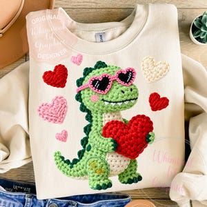 May include: Cream-colored sweatshirt featuring a crocheted green dinosaur wearing heart-shaped sunglasses and holding a red heart. Surrounding the dinosaur are crocheted hearts in red, pink, and cream colors. The text "Original Whimsical Art Graphics Designer" is visible.