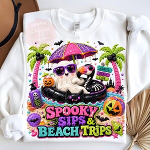 May include: White sweatshirt with a colorful graphic featuring a ghost wearing sunglasses, floating on a black inflatable flamingo, and the text "Spooky Sips & Beach Trips". Includes a pumpkin, palm trees, and bats.