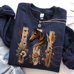 May include: A navy blue sweatshirt with a brown horse and floral design. The design features a horse's head with a flowing mane, surrounded by brown grasses and white flowers. The text "Whimsical Art Graphics" is visible in the bottom right corner.