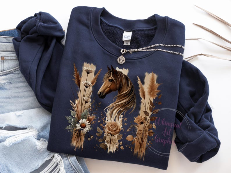 May include: A navy blue sweatshirt with a brown horse and floral design. The design features a horse's head with a flowing mane, surrounded by brown grasses and white flowers. The text "Whimsical Art Graphics" is visible in the bottom right corner.
