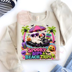 May include: Beige sweatshirt with a colorful graphic featuring a ghost and a skeleton bird in sunglasses, relaxing on a float under a pink umbrella. The text reads "Spooky Sips & Beach Trips". Palm trees, pumpkins, and bats complete the Halloween theme.