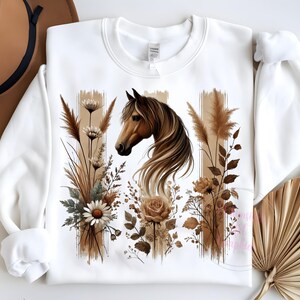 May include: White sweatshirt with a brown horse head and floral design. The horse has a brown mane and tail. The flowers are brown, white, and green. The background is white.