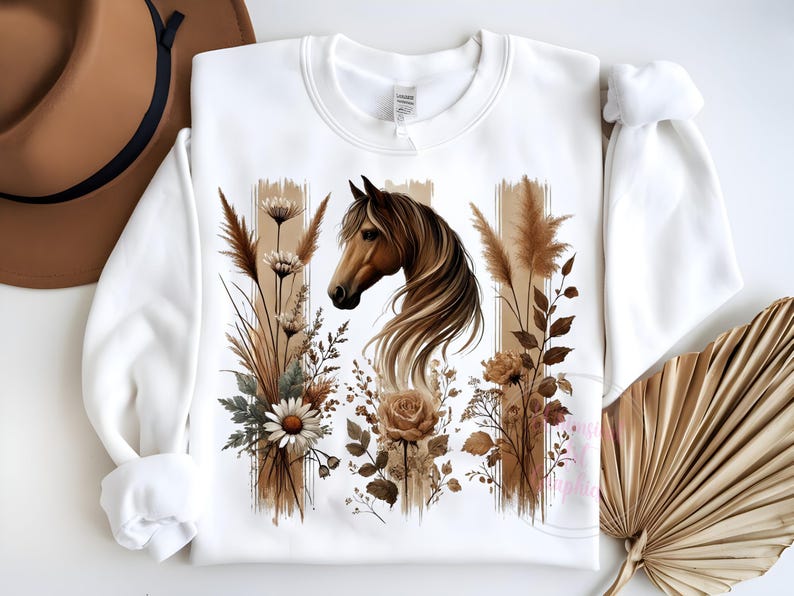 May include: White sweatshirt with a brown horse head and floral design. The horse has a brown mane and tail. The flowers are brown, white, and green. The background is white.