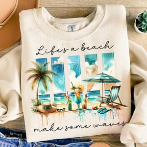 Puede incluir: Una sudadera beige con el texto "Life's a beach make some waves" impreso en ella. El gráfico presenta una escena de playa con palmeras, una sombrilla, sillas de playa y cócteles.