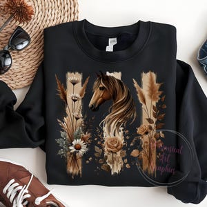 May include: Black sweatshirt with a brown horse and floral design. The horse is surrounded by brown and white flowers and grasses. The design is on a black background with white stripes.