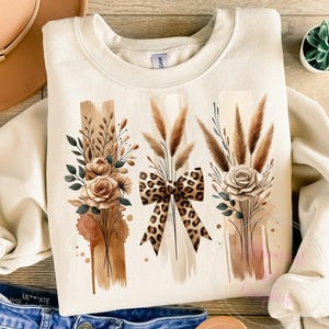 May include: A beige sweatshirt with a graphic design of three bouquets of dried flowers and a leopard print bow. The bouquets are arranged in a vertical row and are surrounded by watercolor-like brown and beige accents.